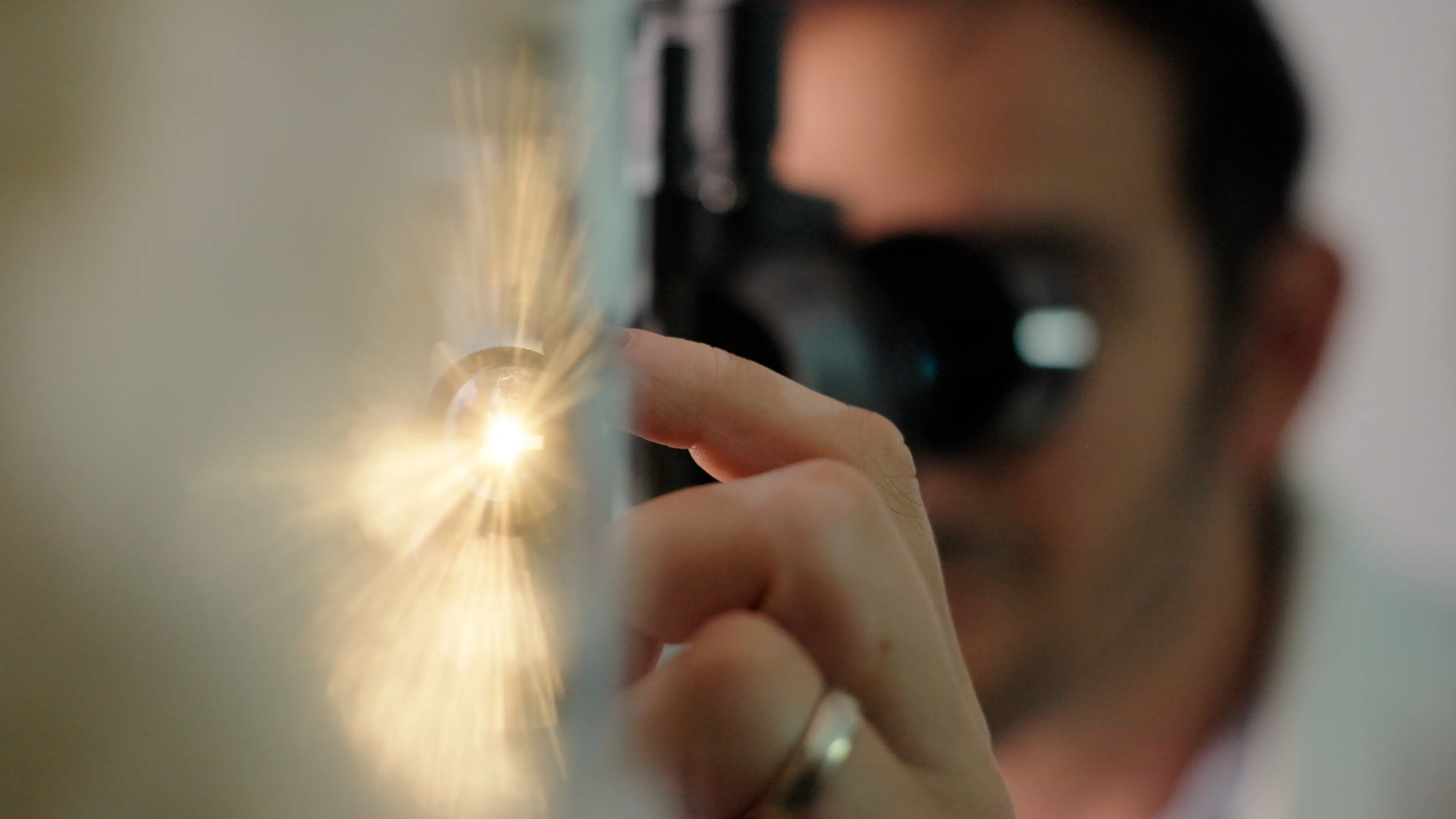 An ophthalmologist using a slit lamp to inspect a patient while a dramatic burst of warm light shines from his lamp on the left side of the image.